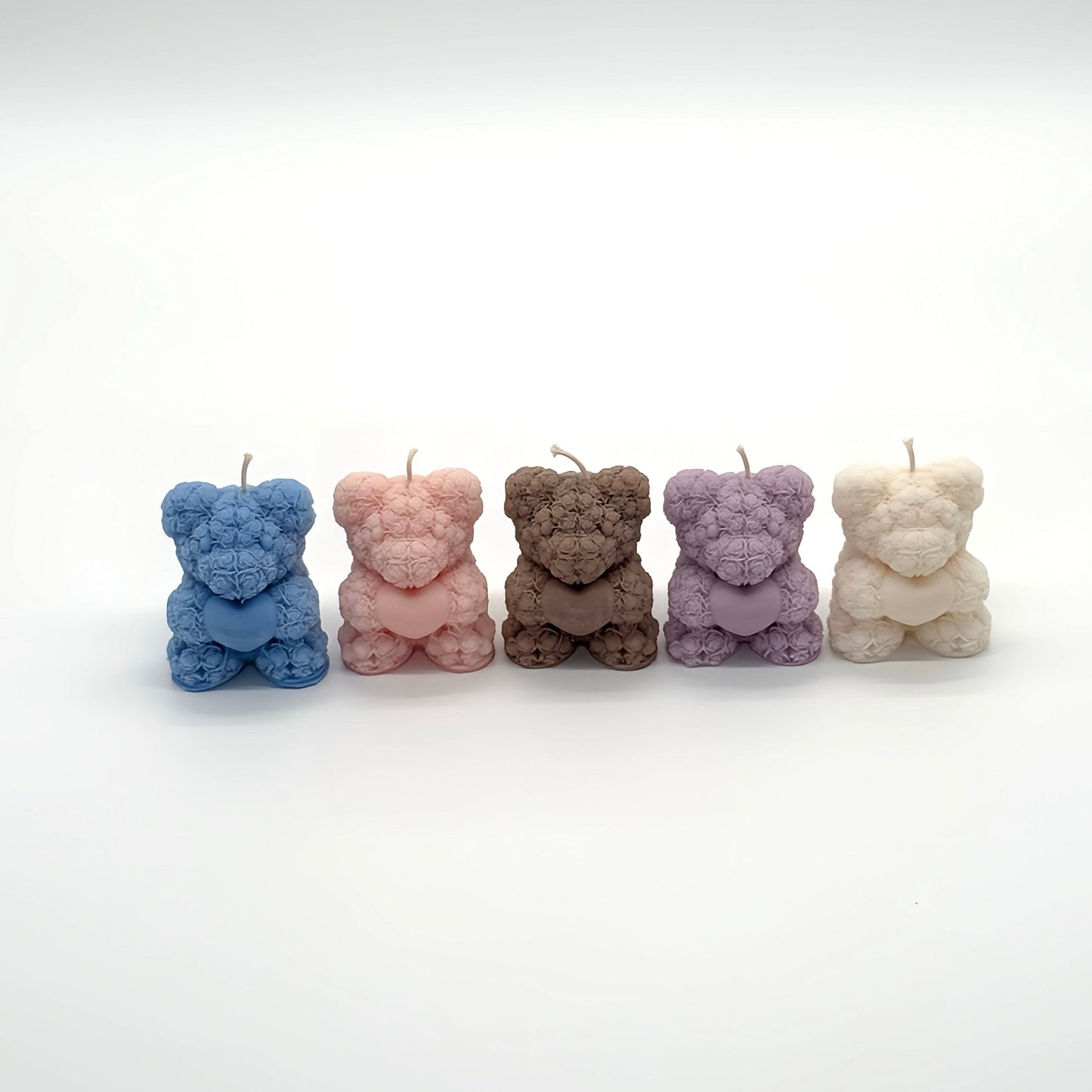 Fünf Teddybär-förmige Kerzen in den Farben Blau, Pink, Lavendel, Braun und Weiß. Jeder Teddy hat ein Rosenblütenmuster und hält ein Herz. Kreative und liebevolle Kerzendesigns auf weißem Studiohintergrund.