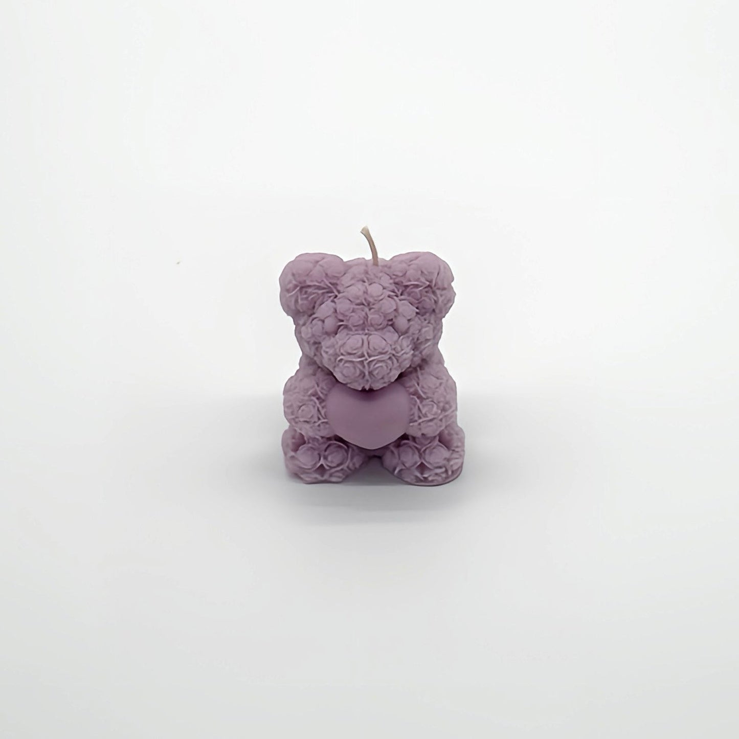 Teddybär-förmige Kerze in Lavendel mit Rosenblütenmuster, hält ein Herz. Sanftes und beruhigendes Kerzendesign für eine entspannte Atmosphäre auf weißem Studiohintergrund.